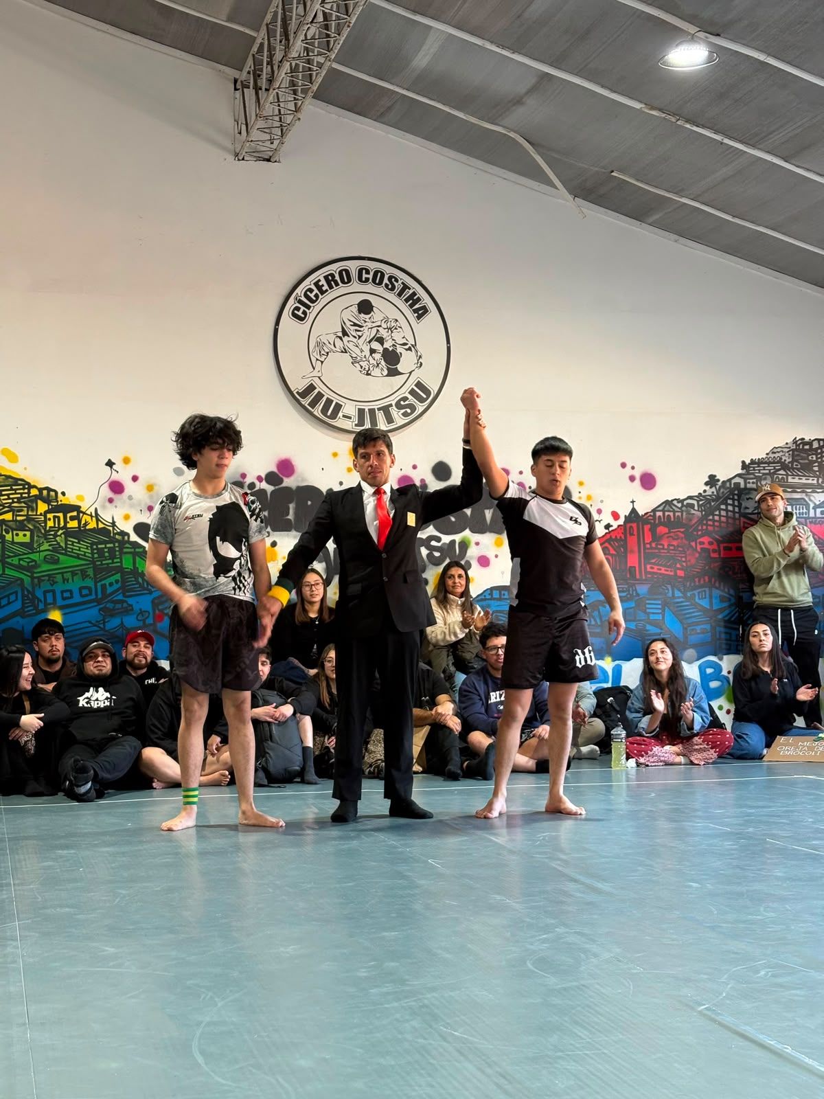 Un alumno con ropa de competición y el brazo levantado junto al árbitro y otro competidor
