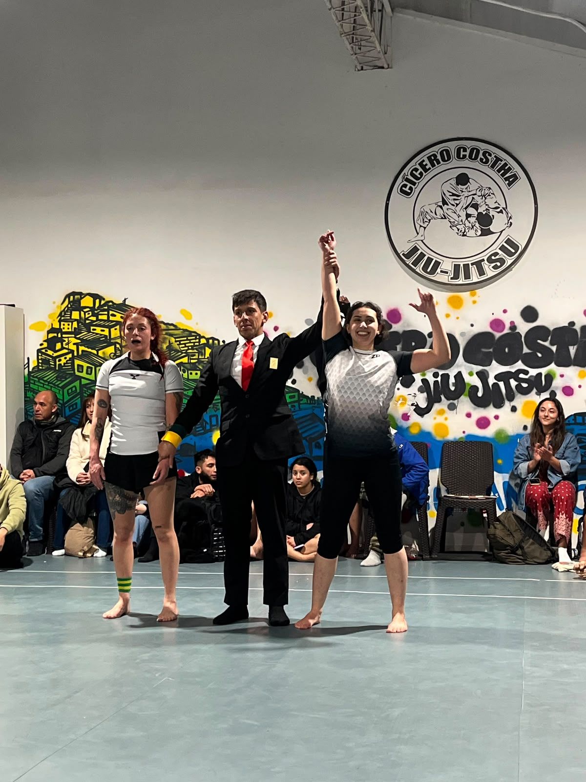 Una alumna con ropa de competición levanta los brazos junto al árbitro y otra competidora en el tatami